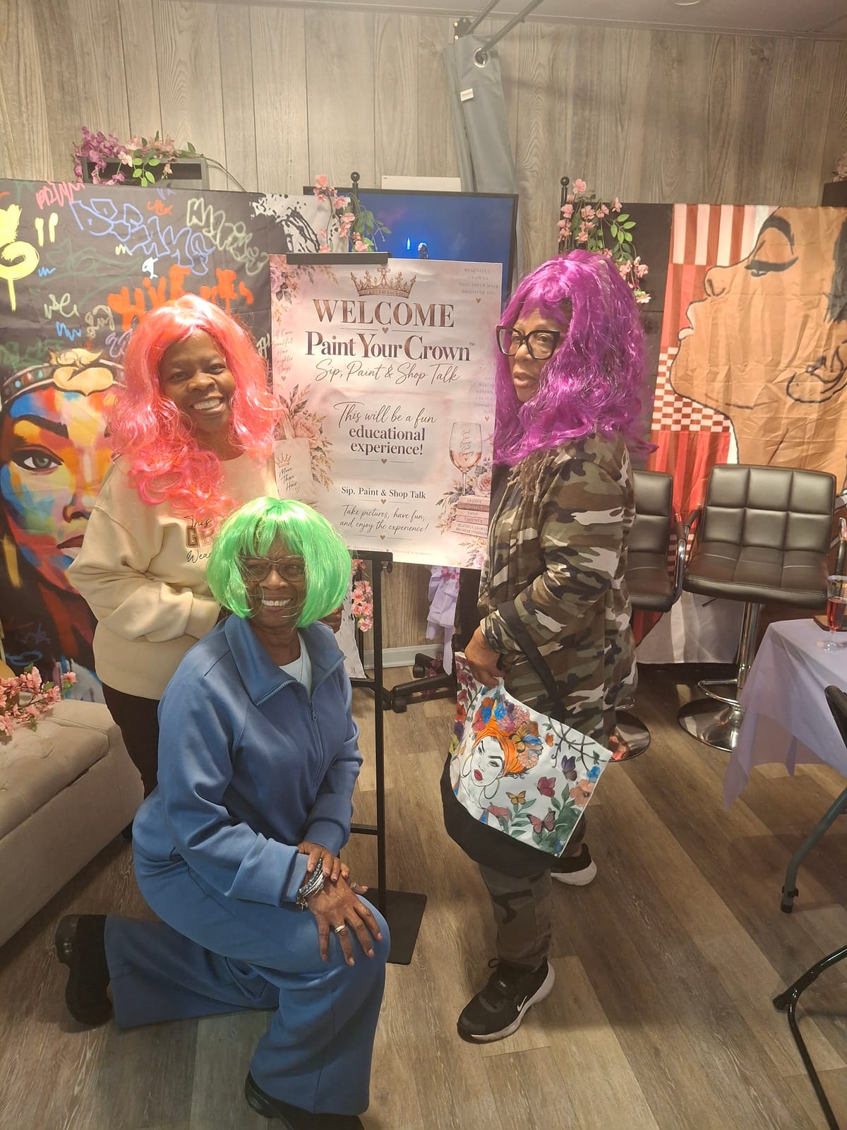 Three guests in bright costume wigs posing beside the Paint Your Crown welcome sign at Natural Touch Hair Studio