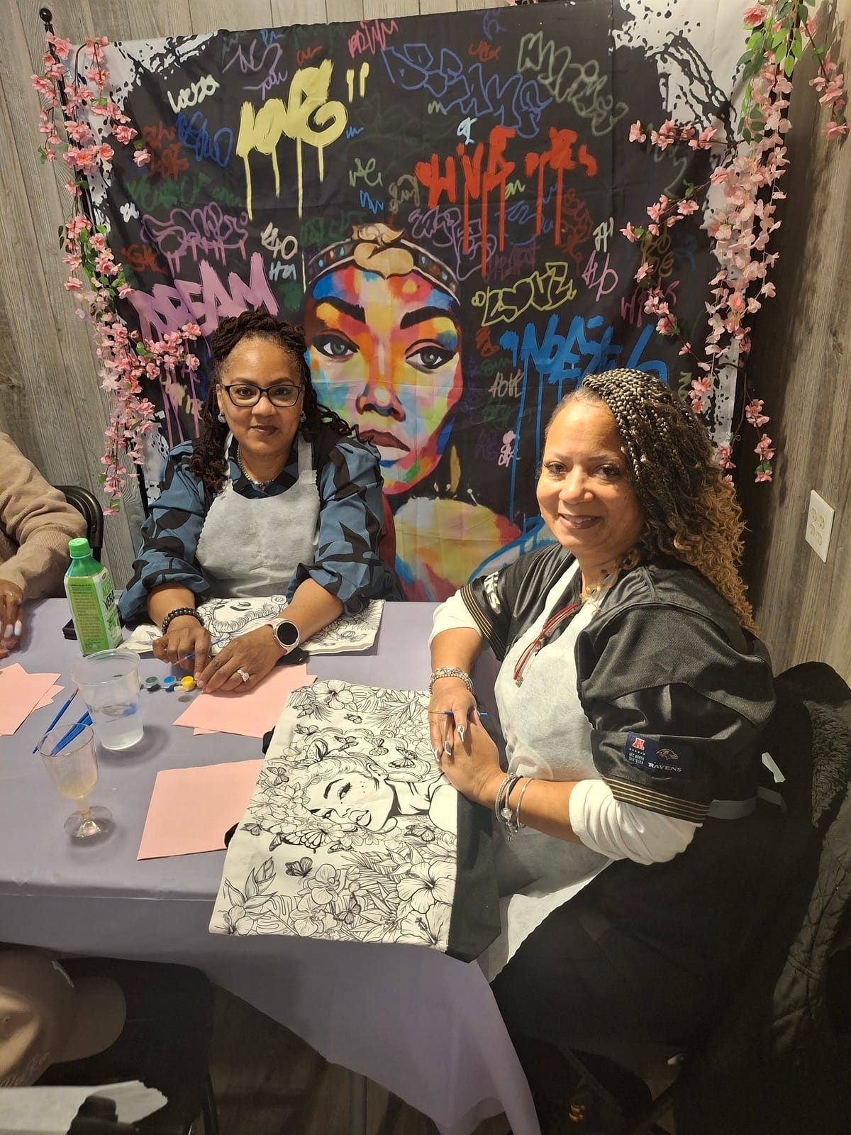 Two guests painting their tote bags in front of the colourful mural backdrop